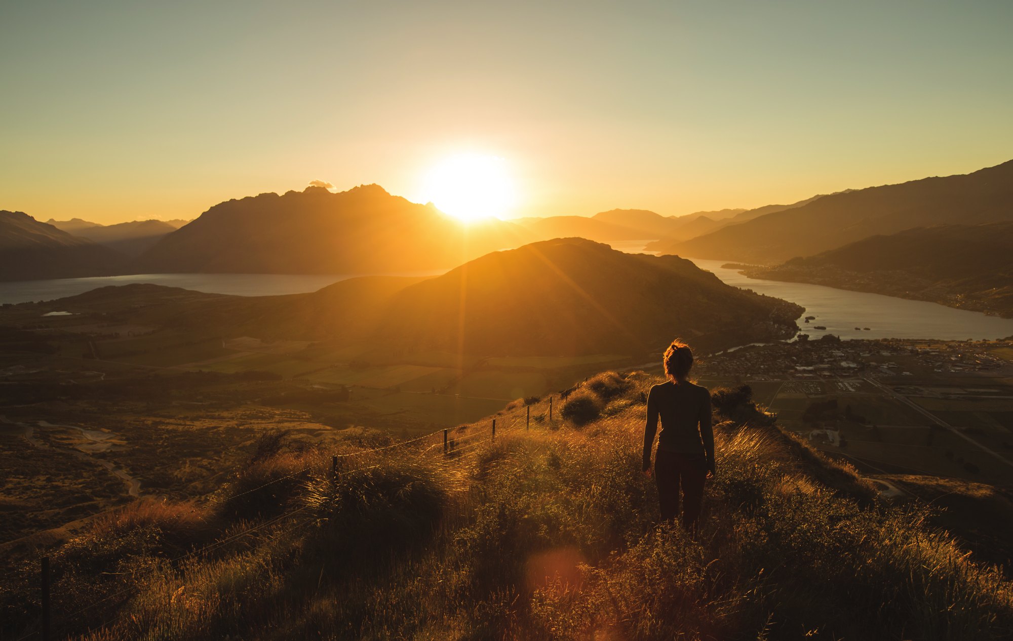 A person standing on a grassy hilltop watching the sun rise over a lake and distant hills